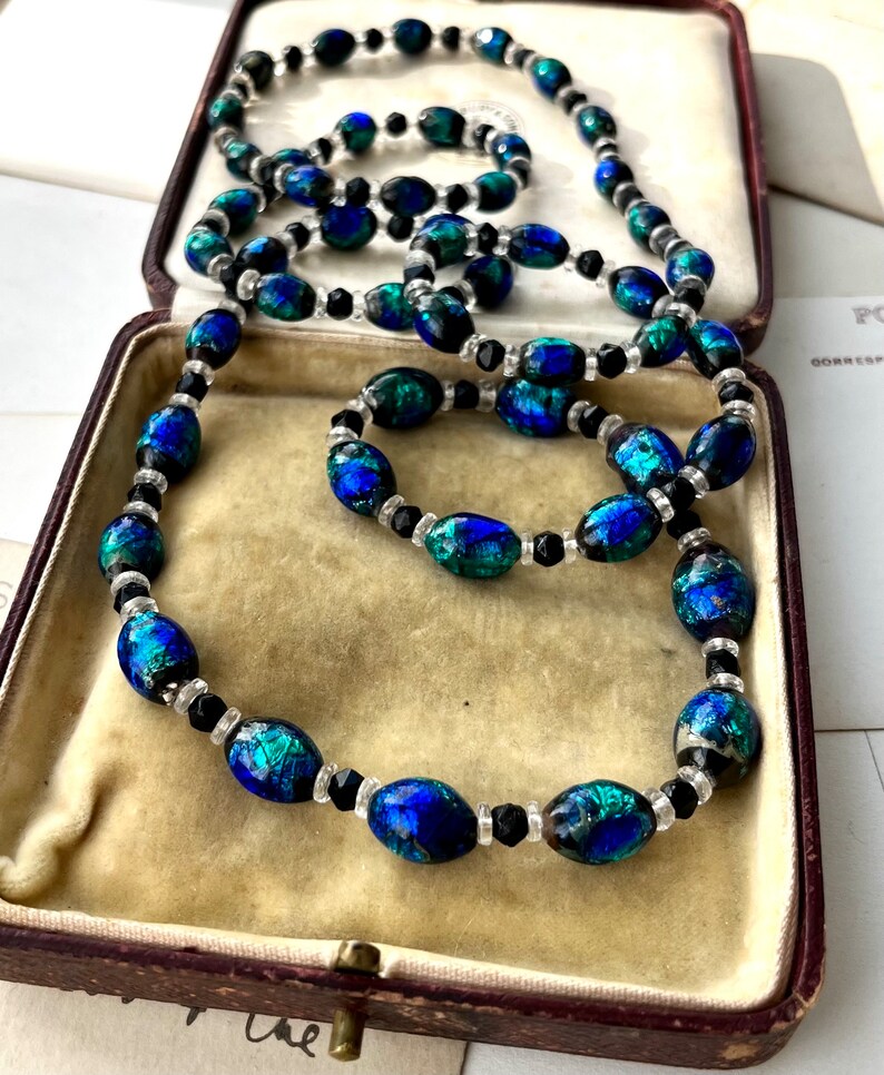 May include: A long necklace and a shorter bracelet made of oval-shaped iridescent blue and green glass beads with black and clear spacer beads.