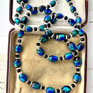 May include: A vintage necklace with oval-shaped iridescent blue and green glass beads interspersed with black and clear round beads. The necklace is displayed in a velvet-lined jewelry box.