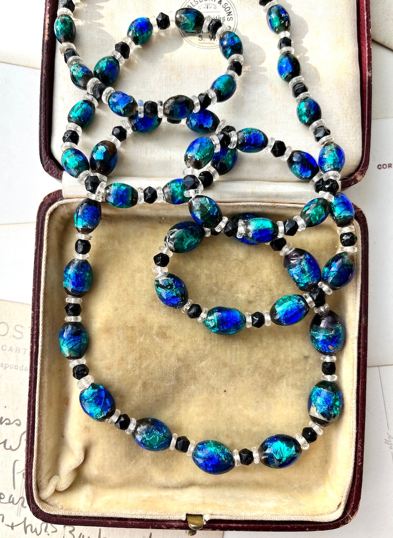 May include: A vintage necklace with oval-shaped iridescent blue and green glass beads interspersed with black and clear round beads. The necklace is displayed in a velvet-lined jewelry box.