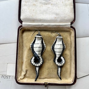 May include: A pair of black and white ceramic snake earrings in a vintage jewellery box. The earrings are in the shape of a snake with a black and white striped pattern. The box is lined with white velvet and has a brown leather exterior. The box has a label that reads "R. Salisbury & Sons Goldsmiths 510 Brixton S.W."