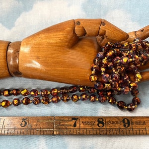 May include: A long strand of iridescent glass beads in shades of purple, gold, and brown. The beads are strung on a clear thread and are approximately 1/2 inch in diameter. The beads are displayed on a wooden hand.