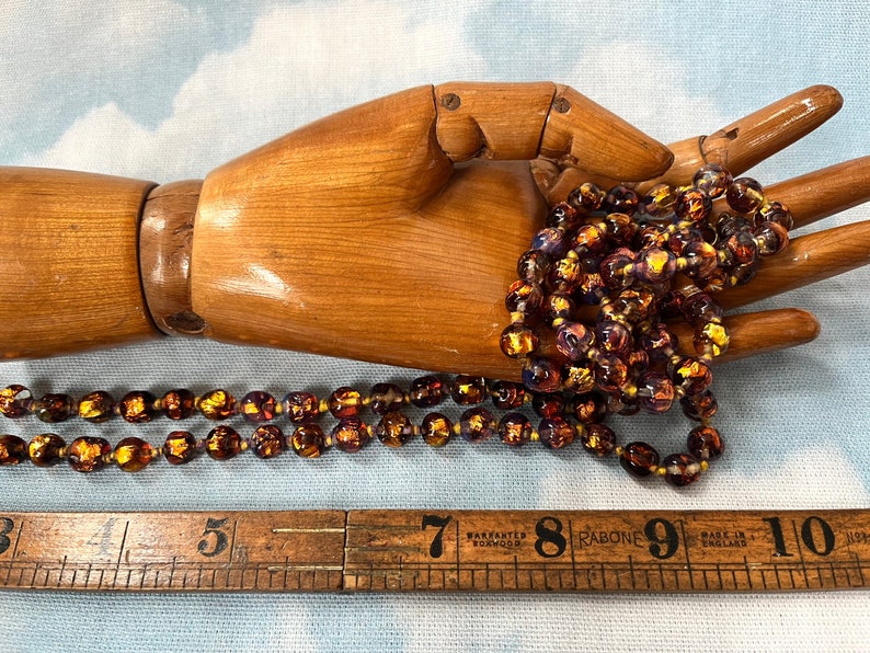 May include: A long strand of iridescent glass beads in shades of purple, gold, and brown. The beads are strung on a clear thread and are approximately 1/2 inch in diameter. The beads are displayed on a wooden hand.