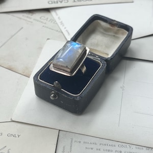 Può includere: Un anello d'argento con una grande pietra di luna rettangolare incastonata in una montatura a castone. L'anello è in una piccola scatola per anelli in velluto nero.