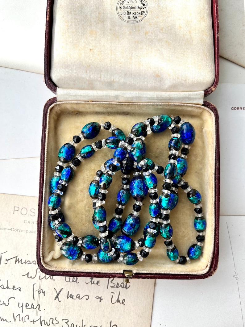 May include: A vintage necklace made of blue and green iridescent glass beads with black and clear spacer beads. The necklace is in a red velvet jewelry box.