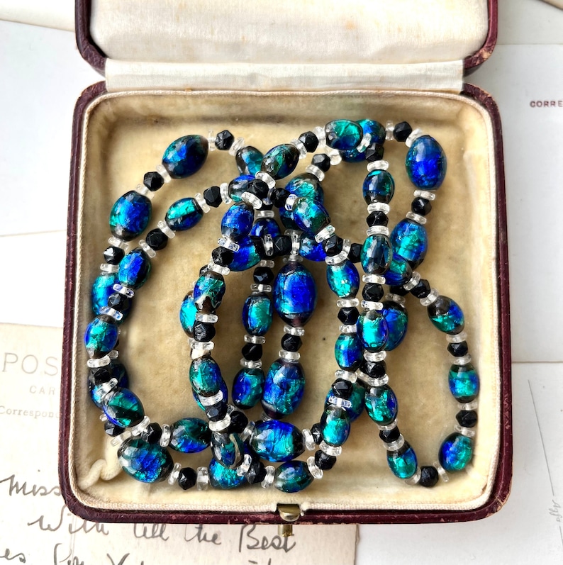 May include: A long necklace made of oval-shaped iridescent blue and green glass beads with black and clear glass beads interspersed throughout. The necklace is displayed in a red velvet jewelry box.