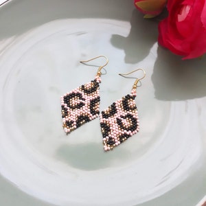 May include: A pair of diamond-shaped earrings with a leopard print design. The earrings feature a pattern of black, pink, and gold beads. The earrings hang from gold-colored ear wires. The earrings are displayed on a light blue plate.