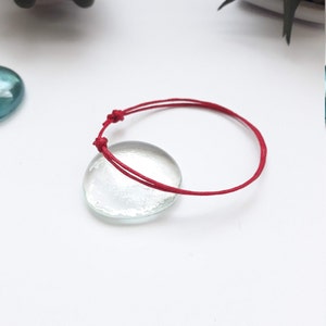 May include: A simple red string bracelet with a knotted closure. The bracelet is lying on a white surface.