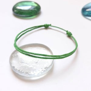 Puede incluir: Un brazalete de cuerda verde con un cierre simple de nudo. El brazalete está sobre una superficie de vidrio transparente.