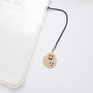 May include: A cream-colored beaded phone charm with a black cord and silver-tone clasp. The charm is in the shape of a small, round face with pink, black, and red bead accents. A clear phone case is visible in the background.