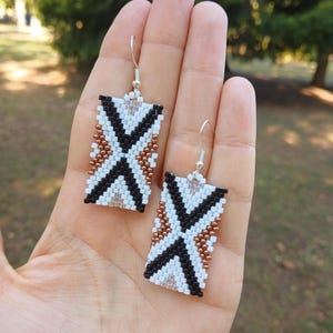 May include: A pair of geometric beaded earrings with a white, black, and copper color scheme. The earrings are rectangular in shape and feature a geometric pattern.