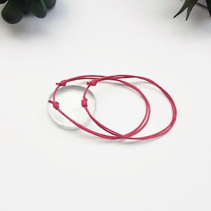 Puede incluir: Dos pulseras de cuerda roja atadas juntas en un nudo. Las pulseras están sobre una superficie blanca.