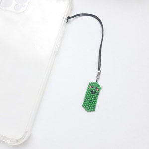 May include: A green beaded phone charm with a smiling face and pink cheeks, attached to a black string.