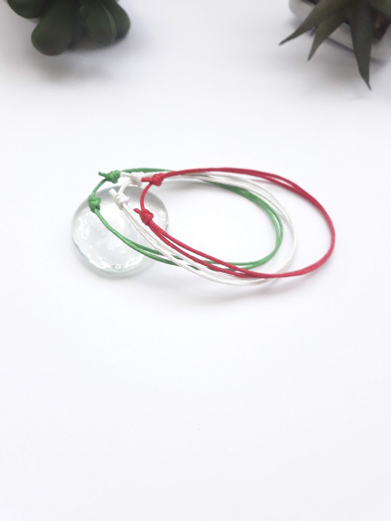 May include: Three friendship bracelets in red, white, and green colors. The bracelets are made of thin cord and are tied together in a knot.