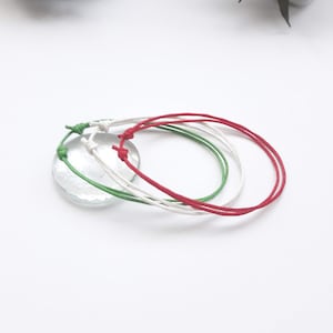 May include: Three colorful friendship bracelets in red, white, and green. The bracelets are tied together and are on a white surface.