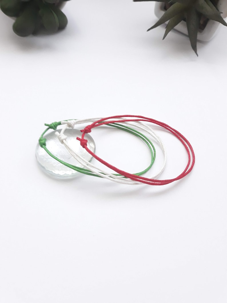May include: Three colorful friendship bracelets in red, white, and green. The bracelets are tied together and are on a white surface.