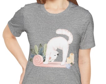 Peut inclure: Un t-shirt gris avec un chat blanc faisant du yoga sur un tapis rose. Le chat est en position de chien tête en bas. Il y a des bougies et des fleurs sur le tapis.