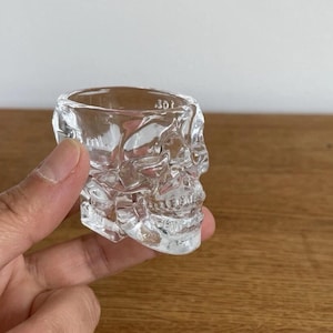 May include: A clear, skull-shaped shot glass. The glass is transparent, allowing light to pass through, and features detailed facial features. The shot glass is held in a hand against a neutral background.