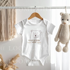 Puede incluir: Un body blanco para bebé con un gráfico de oso y el texto "YOUR DESIGN HERE" cuelga de una percha de madera. Un juguete de oso de peluche de ganchillo cuelga a la derecha. Un tapiz de macramé está en el fondo.