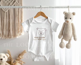 May include: A white baby onesie with a bear graphic and the text "YOUR DESIGN HERE" is hanging on a wooden hanger. A crocheted teddy bear toy hangs to the right. A macrame wall hanging is in the background.