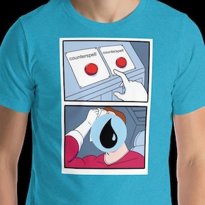 May include: A light blue t-shirt featuring a comic strip. The comic shows two panels: the top panel has two buttons labeled "counterspell", and the bottom panel shows a person sweating with a black teardrop over their face.