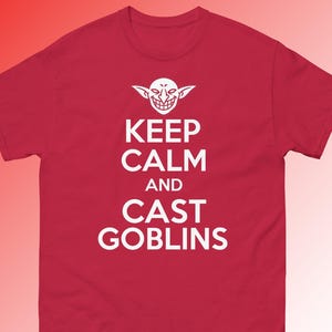 May include: A red short-sleeved t-shirt featuring a white graphic of a grinning goblin face above the text "KEEP CALM AND CAST GOBLINS" printed in a bold, sans-serif font centred on the chest of the garment.