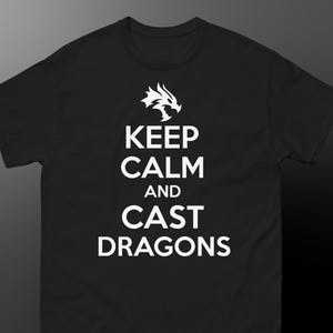 May include: Black t-shirt featuring the white text "KEEP CALM AND CAST DRAGONS" and a white dragon graphic. The shirt has a crew neck and short sleeves, suitable for casual wear.