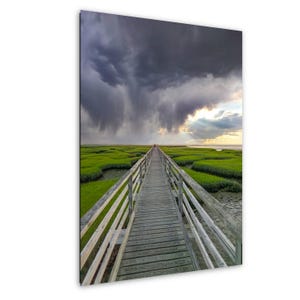 May include: A wooden boardwalk extends into a green marsh under a dramatic sky. Dark storm clouds loom overhead, with rain falling in the distance. The boardwalk leads to a distant figure, with sunlight breaking through the clouds.