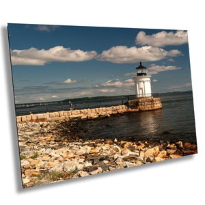 May include: A white lighthouse with a black lantern room stands at the end of a stone breakwater extending into dark blue water. The foreground features a rocky shoreline under a bright blue sky with scattered white clouds and a person walking.