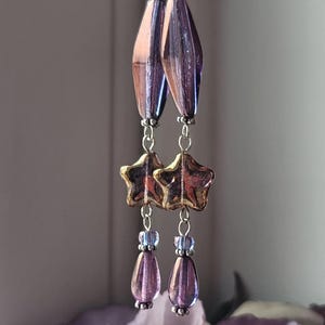 May include: A pair of dangling earrings with a celestial theme. The earrings feature silver hooks, faceted purple and bronze-colored glass beads, gold star-shaped charms, and teardrop-shaped purple beads. The earrings are suspended in front of a blurred background.