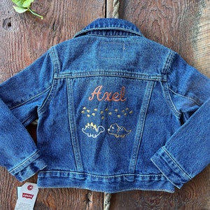 May include: A blue denim jacket with the name "Axel" embroidered on the back in orange thread. Two small yellow dinosaurs are embroidered below the name. A Levi's tag is attached to the bottom left corner.