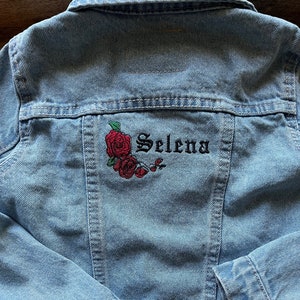 May include: Light blue denim jacket with embroidered red roses and the name "Selena" in black script. The jacket has a classic design with a Levi's tag attached.