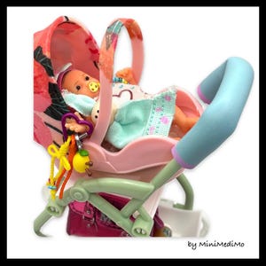 May include: A miniature baby doll in a pink stroller with a floral patterned canopy. The stroller has a light blue handle and a colourful toy attached. The baby is wearing a dummy and a light blue and white outfit.