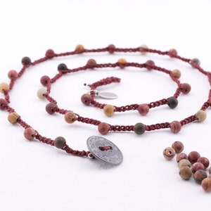 May include: A red cord necklace with brown, green, and yellow stone beads. The necklace has a silver charm and a silver coin pendant. There are extra beads scattered on the white surface.
