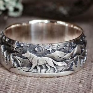 May include: Silver-toned ring featuring a detailed wolf design set against a mountain and starry night scene. The ring's band is engraved with a wolf, trees, mountains, and stars. The ring is a unique piece of jewelry.