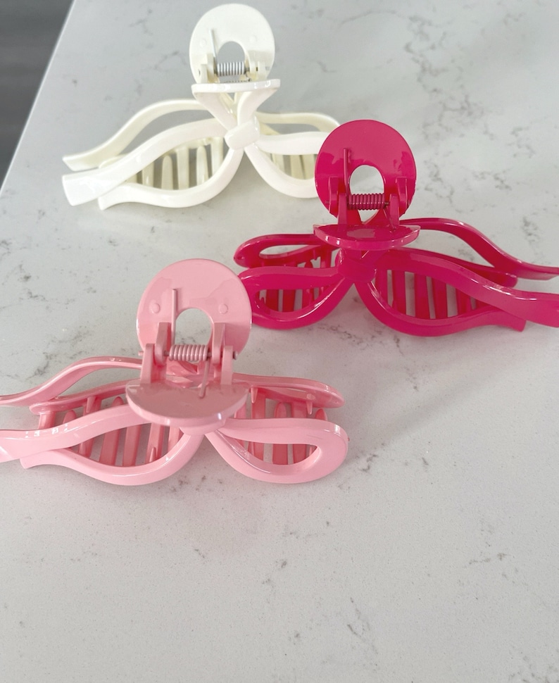 Op de afbeelding: Drie haarclips in verschillende kleuren: wit, roze en felroze. De clips zijn boogvormig en hebben een veermechanisme.