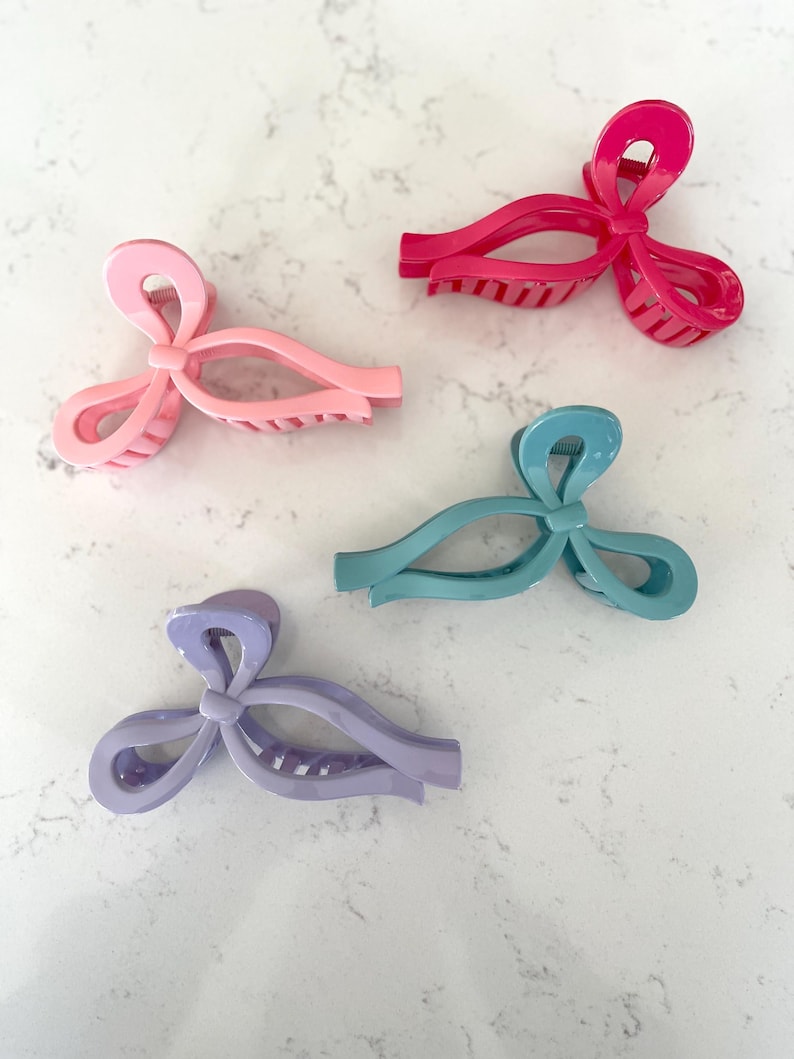 May include: Four plastic hair clips in the shape of bows. The clips are in pink, purple, teal, and red.
