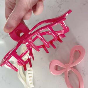 Op de afbeelding: Twee roze plastic haarclips. Een is een grote klauwclip met een ronde basis en meerdere tanden. De andere is een kleinere clip in de vorm van een strik.