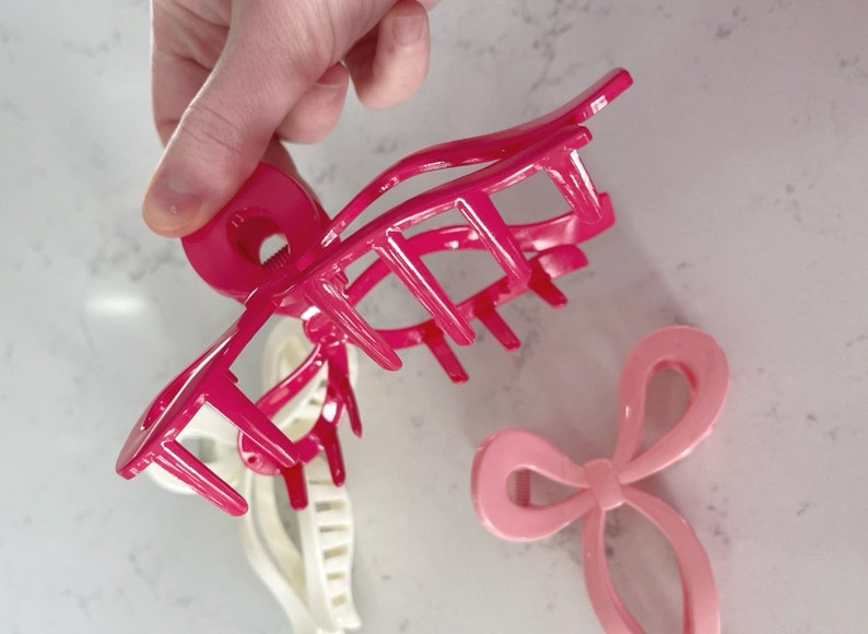 Op de afbeelding: Twee roze plastic haarclips. Een is een grote klauwclip met een ronde basis en meerdere tanden. De andere is een kleinere clip in de vorm van een strik.