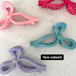 Op de afbeelding: Vier plastic haarclips in verschillende kleuren: roze, paars, teal en felroze. Elke clip heeft de vorm van een strik en een textuur.