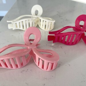 Op de afbeelding: Drie plastic haarclips in roze en wit. De clips zijn gevormd als strikken met een kamvormig ontwerp aan de achterkant.