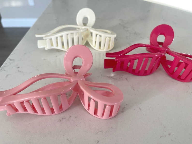 Op de afbeelding: Drie plastic haarclips in roze en wit. De clips zijn gevormd als strikken met een kamvormig ontwerp aan de achterkant.