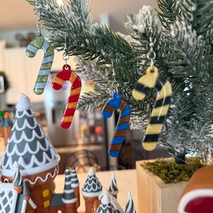 May include: A close-up of a Christmas tree decorated with candy cane earrings in various colors. The earrings are red, gold, blue, and green with silver hooks. A gingerbread house is in the foreground.