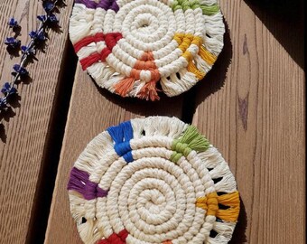 May include: Two handmade macrame coasters with a spiral design. Each coaster features a cream-colored base with colorful accents in red, orange, yellow, green, blue, and purple. A teal mug sits nearby on a wooden surface.