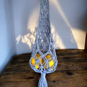 May include: A gray macrame fruit hammock with a wooden ring and tassel, holding yellow lemons. The text "Style Straight" is in red at the top. The hammock hangs on a wooden table.