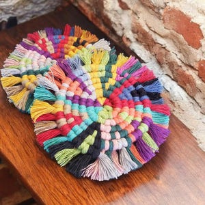 May include: Two colorful round crocheted coasters with fringe. The coasters are made with a variety of colors, including red, orange, yellow, green, blue, purple, and black.