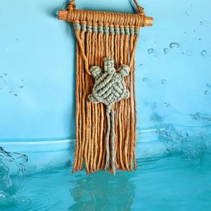 May include: A macrame wall hanging with a blue turtle design. The hanging is made with natural colored yarn and has a wooden dowel at the top.