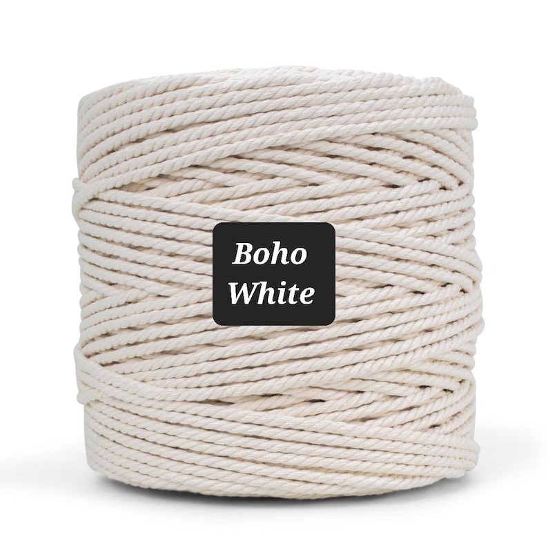 May include: A tightly wound spool of off-white macrame cord. The cord is thick and has a twisted texture. A black square label with the words "Boho White" in white lettering is affixed to the front of the spool. Ideal for crafting.