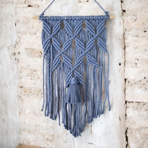 May include: A blue macrame wall hanging with a geometric diamond pattern and a tassel. The wall decor is suspended from a wooden dowel and a string. The background is a textured, light-colored brick wall.