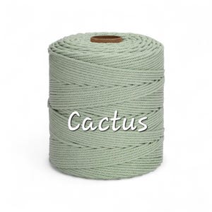 May include: A cylindrical spool of light sage green macrame cord featuring a twisted rope texture. The word Cactus is printed in white script across the center of the spool, which has a brown cardboard core visible at the top center.