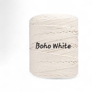 May include: A cylindrical spool of off-white macrame cord wound in a continuous spiral pattern. The text "Boho White" is printed in black, sans-serif lettering across the center of the cord spool, which features a textured, braided surface.